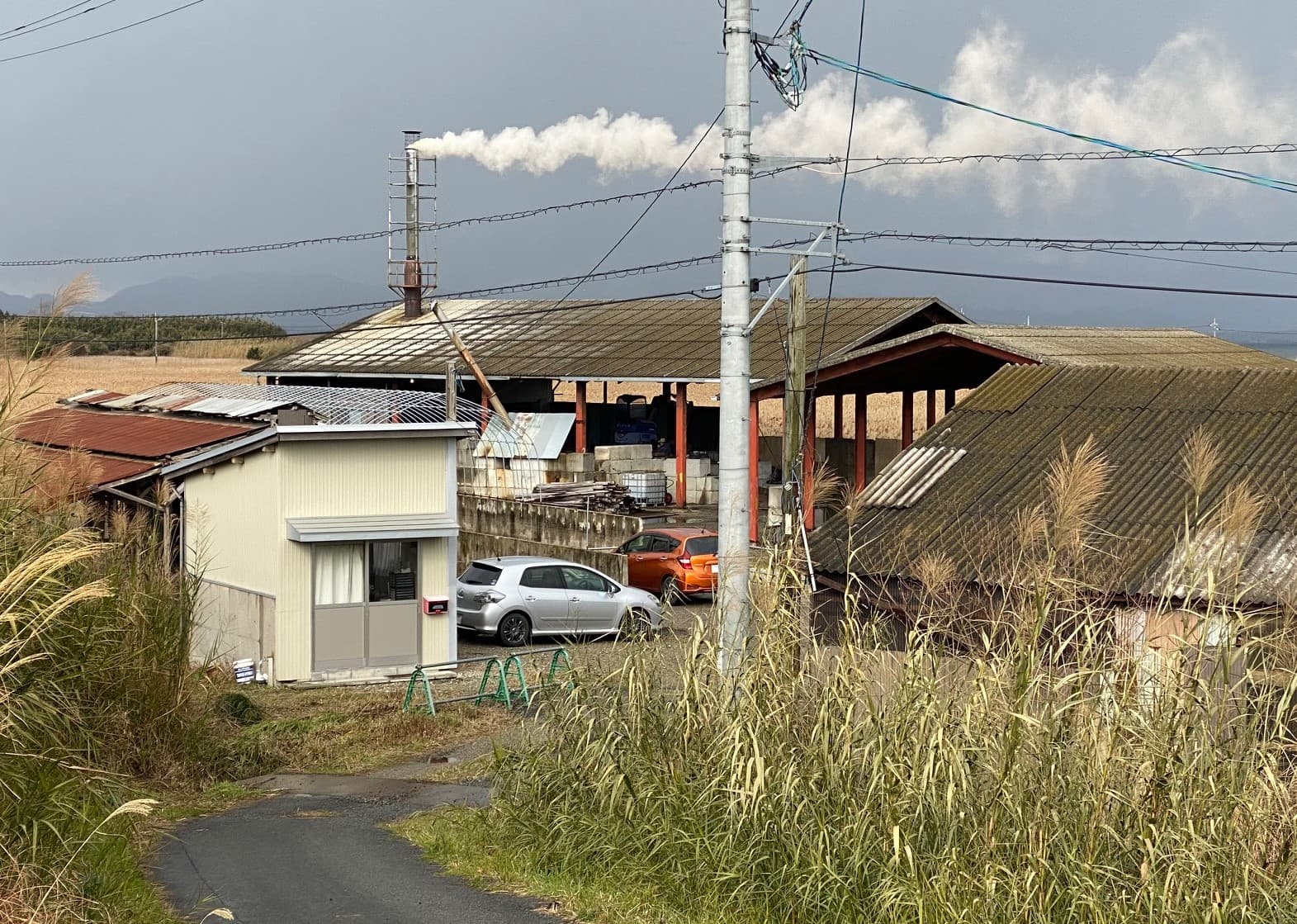 IZUTAN の工場全景。煙突から白煙が立ち、ススキの草地と山が広がる山陰の風景。
